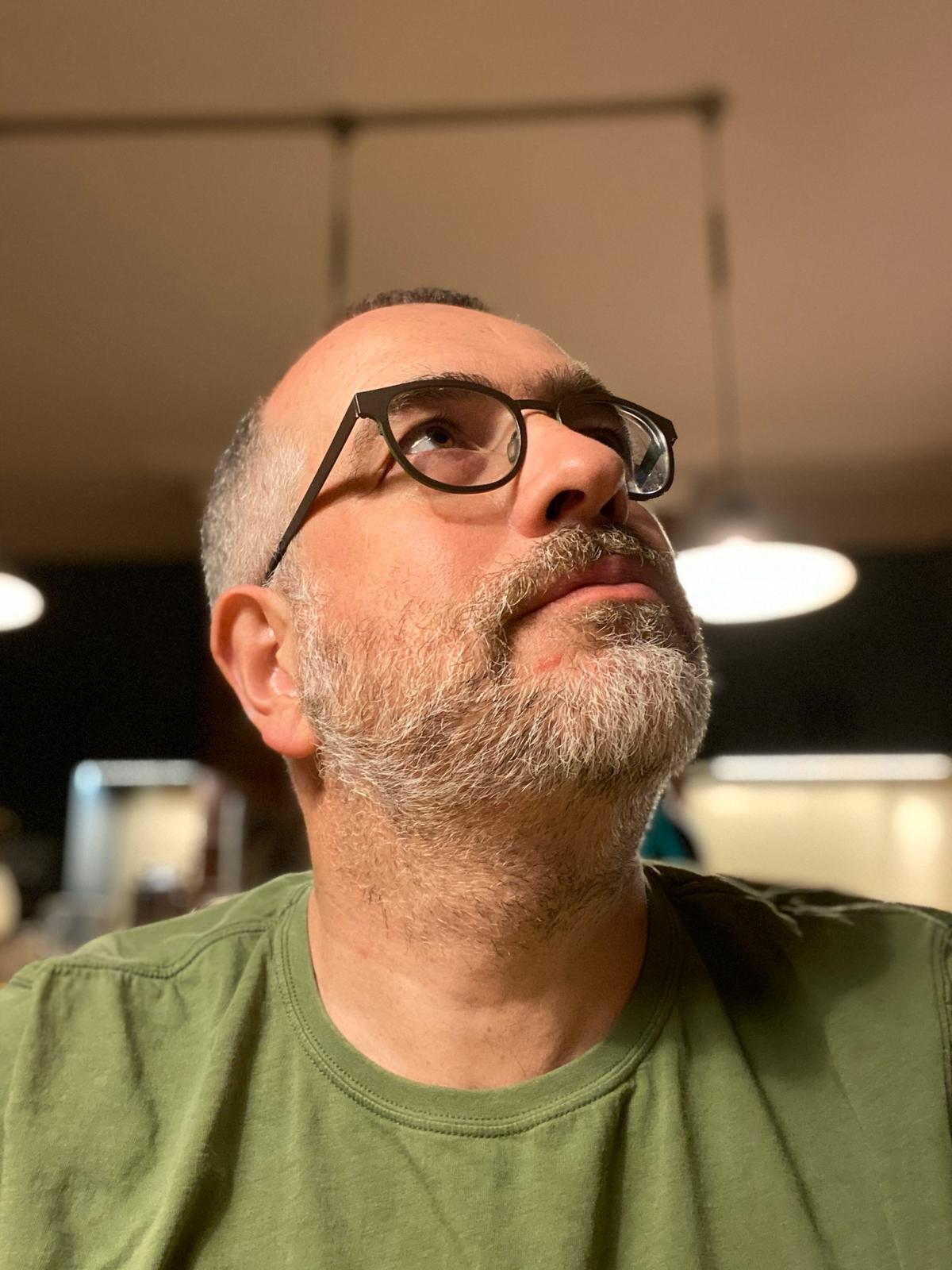 A man with greying, close-cropped hair looks up and to the right. He wears glasses and a green t-shirt.